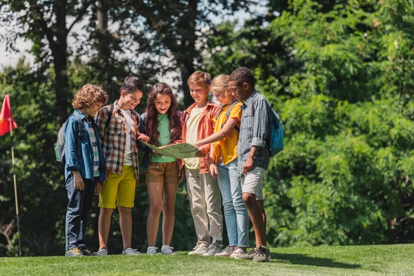 Happy group of multicultural kids looking at map near trees in park — Stock Photo