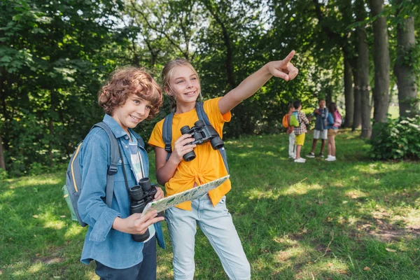 Selective focus of happy kid with binoculars pointing with finger near friend holding map — Stock Photo