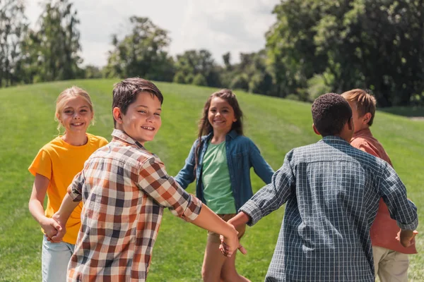 Messa a fuoco selettiva di allegri bambini multiculturali che si tengono per mano nel parco — Foto stock