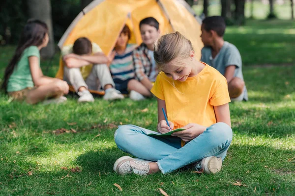 Enfoque selectivo de niño feliz escribiendo en cuaderno cerca de amigos multiculturales en el campamento - foto de stock