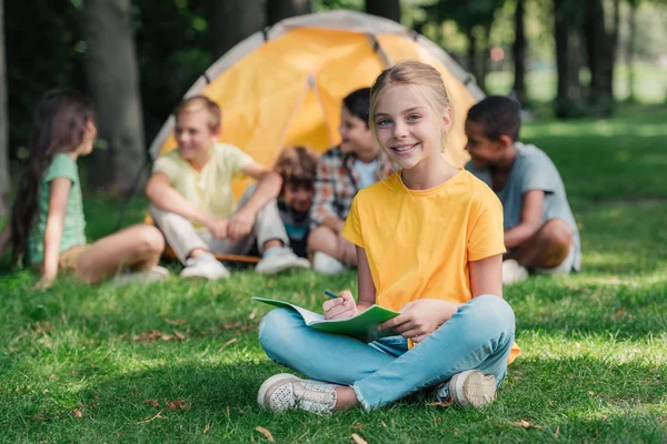 Selective focus of cheerful kid holding notebook near multicultural friends in camp — Stock Photo
