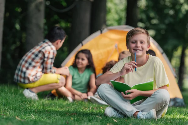 Fuoco selettivo di bambino felice che tiene il notebook e la matita vicino ad amici in campo — Foto stock