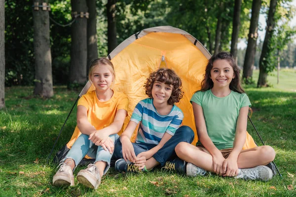 Heureux garçon bouclé assis avec des amis mignons près du camp — Photo de stock