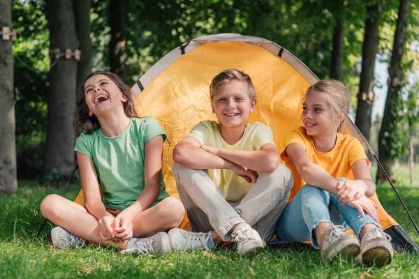 Счастливый мальчик, сидящий с симпатичными друзьями, смеющийся возле лагеря — стоковое фото
