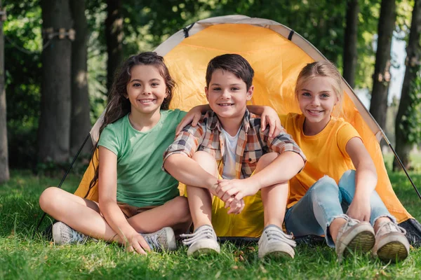 Menino feliz sentado com amigos bonitos perto do acampamento — Fotografia de Stock