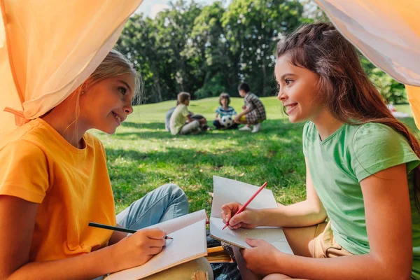 Enfoque selectivo de niños felices escribiendo en cuadernos cerca del campamento - foto de stock