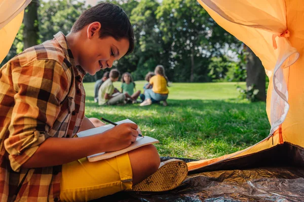 Selektiver Fokus des glücklichen Jungen, der im Lager in Notizbuch schreibt — Stockfoto
