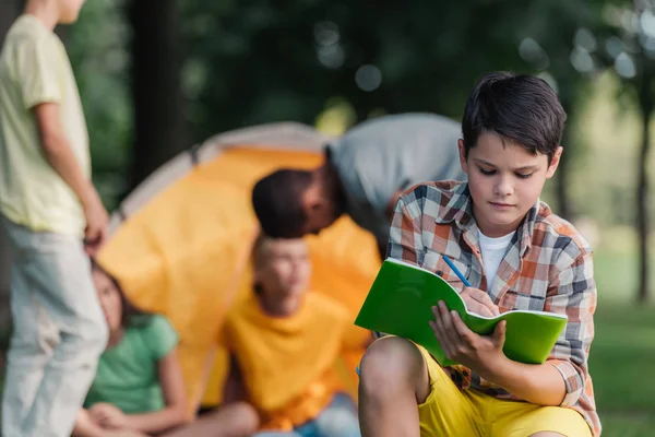 Selective focus of kid holding notebook and pencil near friends in camp — Stock Photo