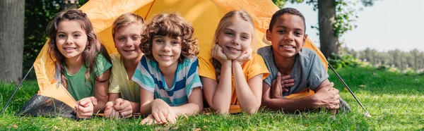 Panoramic shot of happy multicultural kids smiling while lying near camp — Stock Photo