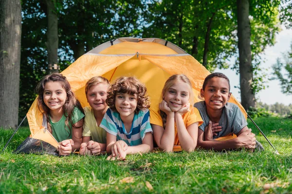 Alegres niños multiculturales sonriendo mientras yacen cerca del campamento - foto de stock