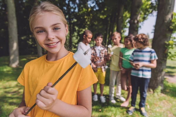 Enfoque selectivo de niño feliz sosteniendo malvavisco dulce en palo cerca de amigos multiculturales - foto de stock