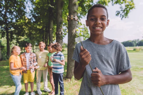 Enfoque selectivo de niño afroamericano feliz sosteniendo malvavisco dulce en palo cerca de amigos - foto de stock