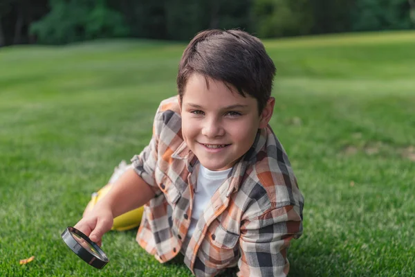 Selective focus of happy boy holding magnifier while lying on grass — Stock Photo