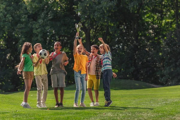 Счастливые мультикультурные дети, стоящие на траве с футболом и трофеем — стоковое фото