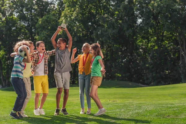 Niños multiculturales felices saltando en la hierba con el fútbol y el trofeo - foto de stock