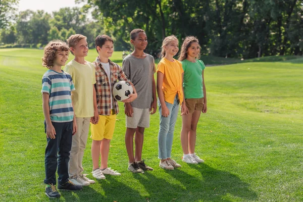 Fröhliche multikulturelle Kinder stehen mit Fußball auf dem Rasen — Stockfoto