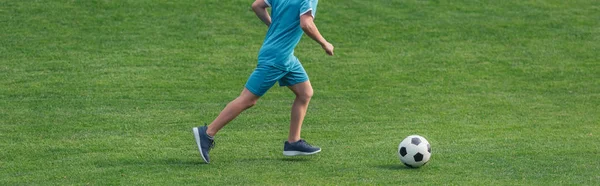 Mierda panorámica de niño en ropa deportiva corriendo sobre hierba verde con el fútbol - foto de stock