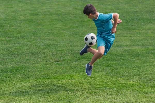 Bambino in abbigliamento sportivo che salta su erba verde con calcio — Foto stock