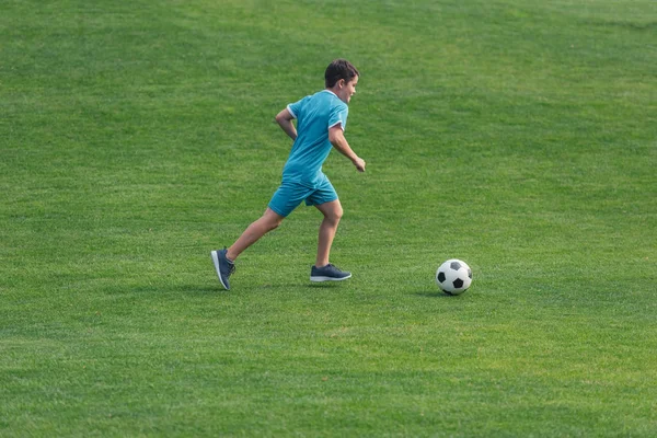 Ребенок в спортивной одежде бегает по зеленой траве с футболом — стоковое фото