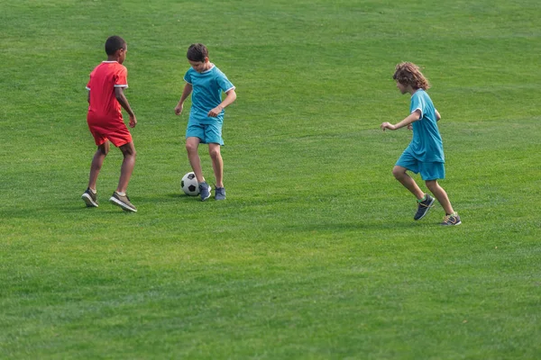 Cute multicultural boys in sportswear playing football on grass — Stock Photo