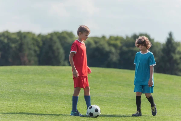 Милый кудрявый мальчик играет в футбол с другом на траве в парке — стоковое фото