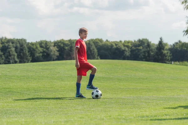 Милый ребенок в спортивной одежде, стоящий на зеленой траве с футболом — стоковое фото