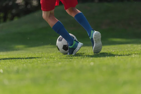 Vista ritagliata del bambino in abbigliamento sportivo in esecuzione su erba verde con calcio — Foto stock