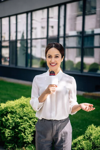 Atractivo periodista en ropa formal sosteniendo micrófono y hablando afuera - foto de stock