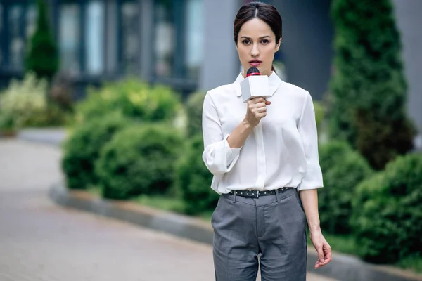 Atractivo periodista en ropa formal sosteniendo micrófono y hablando afuera - foto de stock