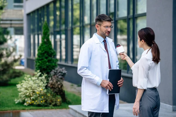 Журналіст тримає мікрофон і розмовляє з красивим лікарем у білому пальто — стокове фото