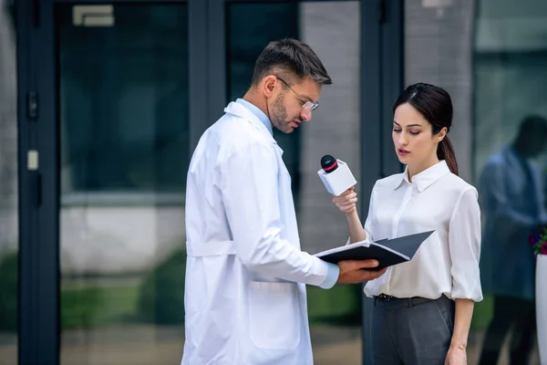 Journaliste tenant un microphone et parlant avec beau médecin en manteau blanc — Photo de stock