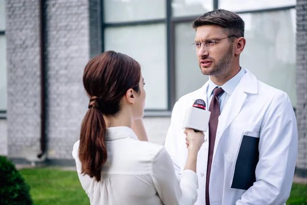 Giornalista che tiene il microfono e parla con un bel dottore in camice bianco — Foto stock