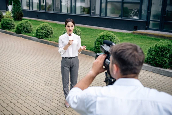 Selective focus of attractive journalist holding microphone and cameraman shooting her outside — Stock Photo