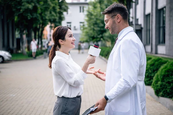 Periodista sosteniendo micrófono y hablando con el guapo doctor en bata blanca - foto de stock