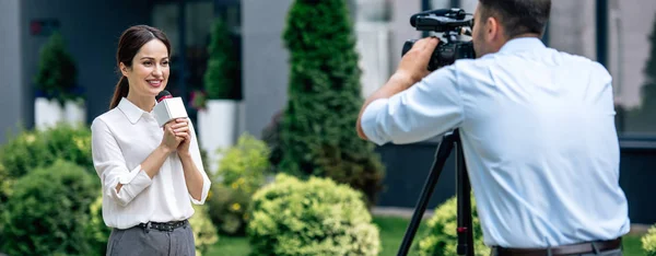 Panoramic shot of attractive journalist holding microphone and cameraman shooting her outside — Stock Photo