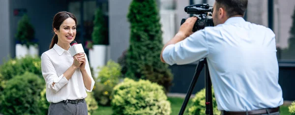 Prise de vue panoramique de journaliste attrayant tenant microphone et caméraman tirant sur elle à l'extérieur — Photo de stock