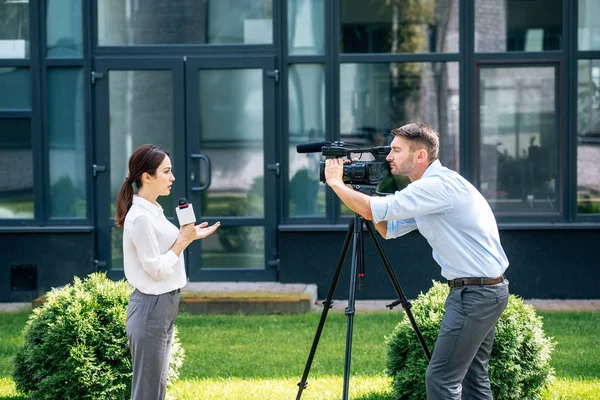 Side view of attractive journalist holding microphone and cameraman shooting her outside — Stock Photo