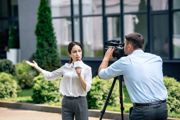 Attractive journalist holding microphone and cameraman shooting her outside — Stock Photo