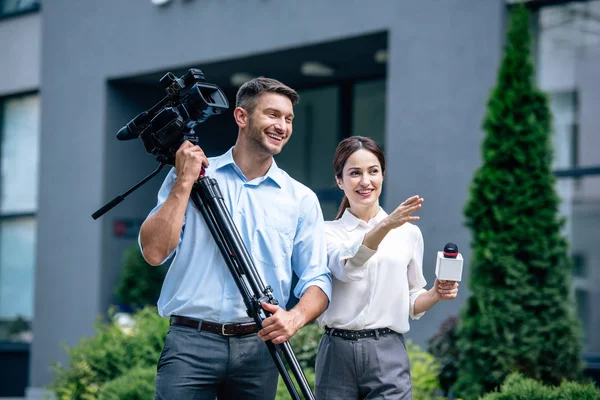Attrayant journaliste tenant microphone et caméraman tenant caméra vidéo numérique — Photo de stock