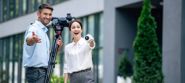 Panoramic shot of attractive journalist holding microphone and cameraman holding digital video camera — Stock Photo