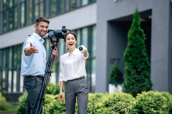 Atractivo periodista sosteniendo micrófono y camarógrafo sosteniendo cámara de video digital - foto de stock