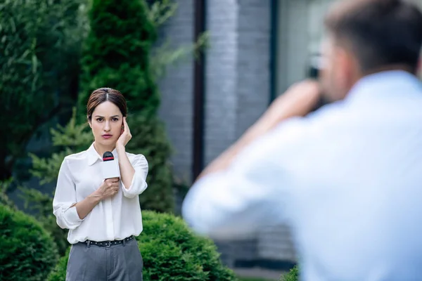 Selective focus of attractive journalist holding microphone and cameraman shooting her outside — Stock Photo