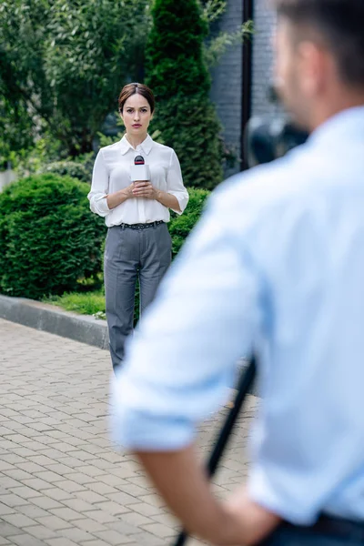 Selective focus of attractive journalist holding microphone and cameraman shooting her outside — Stock Photo