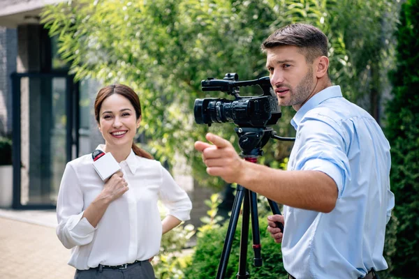 Atractivo periodista sosteniendo micrófono y camarógrafo señalando con el dedo - foto de stock