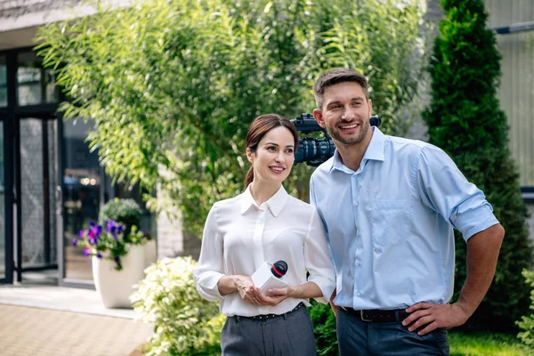 Attraktive Journalistin mit Mikrofon und Kameramann, die lächelt und wegschaut — Stockfoto