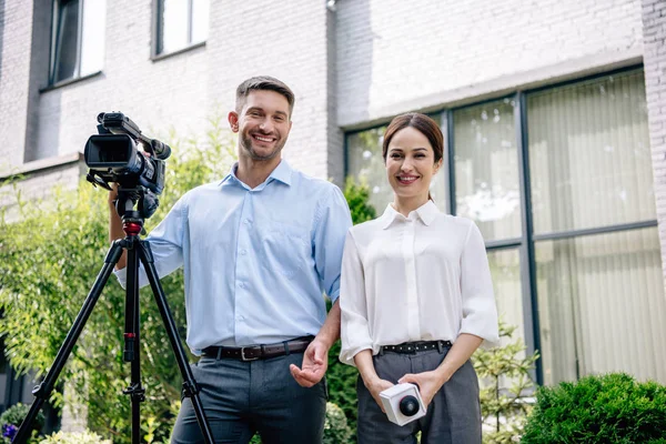 Atractivo periodista sosteniendo micrófono y camarógrafo sosteniendo cámara de video digital - foto de stock