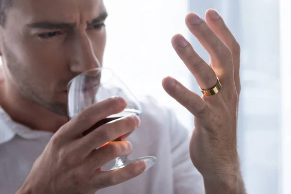 Selective focus of depressed man in shirt drinking cognac and looking at ring at home — Stock Photo