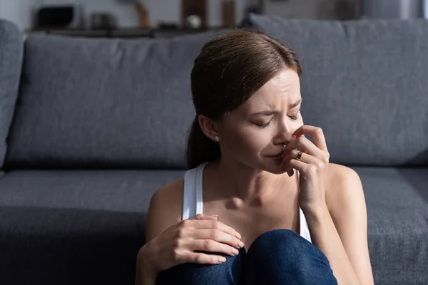 Mujer joven molesta con anillo sentado cerca del sofá y llorando en casa - foto de stock