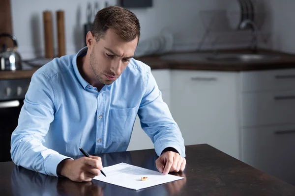 Homme triste en chemise assis à table avec anneau et signer les documents de divorce — Photo de stock