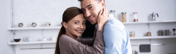 Plano panorámico de pareja sonriente abrazando y mirando a la cámara en casa - foto de stock